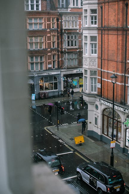 A busy street scene in Marylebone featuring a row of classic brick residential buildings with balconies and large windows. Some buildings are under renovation, with scaffolding covering parts of the facades. Pedestrians are walking along the pavement, including individuals carrying shopping bags and a person pushing a trolley. Street furniture includes a black lamppost with decorative elements, traffic lights showing red and green signals, and a clock mounted on the lamppost. Storefronts on the ground level display signs, one indicating a house removal or moving service with the company name Marylebone Man and Van, situated on a building corner. The sky is partly cloudy, casting diffused natural light over the scene. The image captures the urban environment typical of home relocation logistics, with visible street activity and building features relevant to furniture transport and packing during a house move.