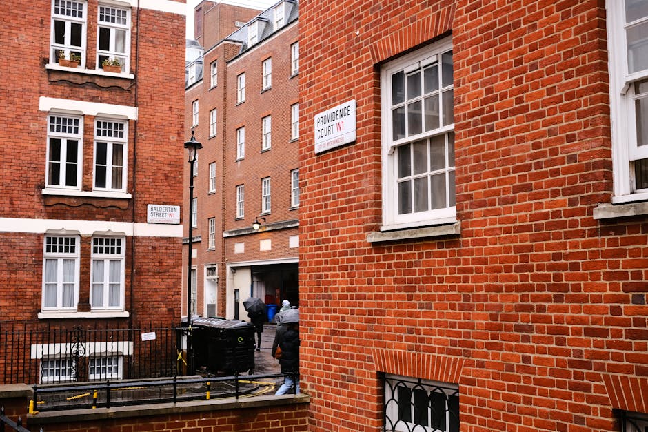 Photograph of a home relocation scene outside a red brick residential building in Marylebone. The image shows two men in casual clothing, one wearing a hooded jacket, lifting a large cardboard box wrapped in plastic wrap towards a black van parked close to the building's entrance. The van is partially visible, with an open rear door indicating active loading. In the background, there are additional apartment windows with white frames, and a narrow alleyway with a black wheelie bin and a street lamp. The scene is lit with natural daylight, and the surrounding environment includes street signs indicating the intersection of Balderton Street and Providence Court in Marylebone, Westminster. The image captures the process of moving and packing items during furniture transport and house removals, with a focus on handling cardboard boxes and strategic loading in an urban setting, as managed by Marylebone Man and Van.