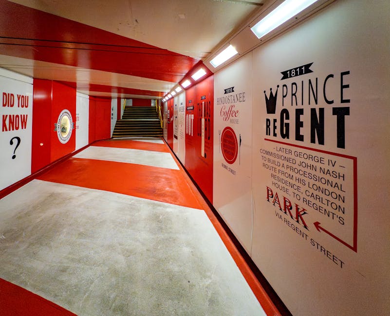 A brightly lit underground corridor with white walls decorated with vintage-inspired red and black signage, including the 'Prince Regent' sign and historical references. The floor features a skid-resistant concrete surface with a central red section leading toward a staircase with black and yellow striped safety markings on the steps. On the left, a large white wall displays a question mark symbol in black, amidst various graphic elements. The corridor is used for transporting household or office furniture and boxes during a home relocation, with the environment suited for personnel from [COMPANY_NAME] to load and unload items onto a nearby commercial van parked outside the building. The hallway's lighting includes overhead fluorescent strips, and the area appears clean and organized for the logistics of furniture transport and packing during a house move or removal service. This internal space facilitates efficient loading and unloading processes as part of a professional removals operation, connected to the services detailed on the [PAGE_TITLE] page.
