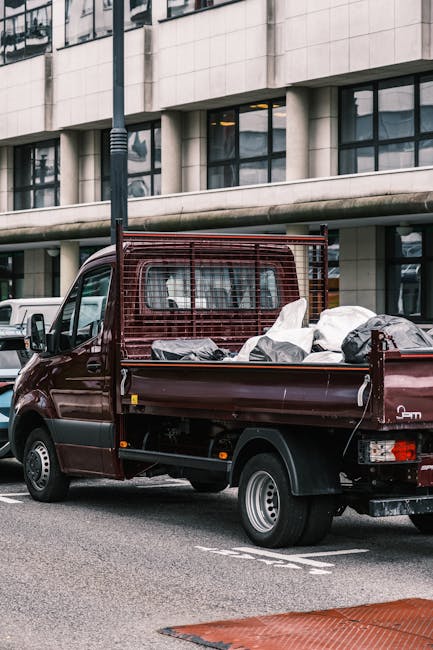 Photograph of a home relocation scene outside a red brick residential building in Marylebone. The image shows two men in casual clothing, one wearing a hooded jacket, lifting a large cardboard box wrapped in plastic wrap towards a black van parked close to the building's entrance. The van is partially visible, with an open rear door indicating active loading. In the background, there are additional apartment windows with white frames, and a narrow alleyway with a black wheelie bin and a street lamp. The scene is lit with natural daylight, and the surrounding environment includes street signs indicating the intersection of Balderton Street and Providence Court in Marylebone, Westminster. The image captures the process of moving and packing items during furniture transport and house removals, with a focus on handling cardboard boxes and strategic loading in an urban setting, as managed by Marylebone Man and Van.