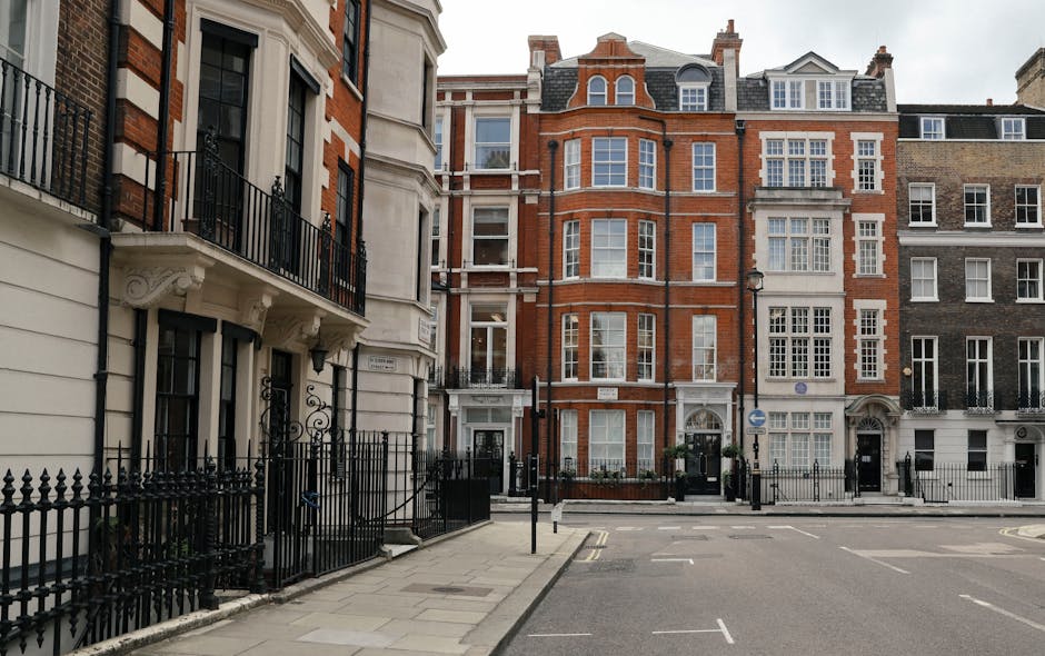 The image shows a quiet Marylebone residential street lined with multi-storey Victorian and Edwardian terraced houses featuring large sash windows, brickwork, and decorative stonework. The street pavement is visible in the foreground, with a black wrought iron fence bordering the front of a white-painted building on the left. The street is empty of pedestrians and vehicles, with a few road markings and parking restrictions visible on the road. The scene is captured during the daytime with natural lighting, highlighting the architectural details of the buildings. This setting exemplifies typical London property facades, often involved in home relocations where furniture and belongings are transported through such urban environments. At [COMPANY_NAME], house removal services are tailored to facilitate efficient furniture transport and packing logistics in areas like Marylebone, ensuring the smooth loading and unloading process for residents moving within or out of these compact homes.