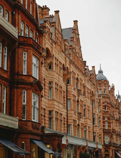The image shows a quiet Marylebone residential street lined with multi-storey Victorian and Edwardian terraced houses featuring large sash windows, brickwork, and decorative stonework. The street pavement is visible in the foreground, with a black wrought iron fence bordering the front of a white-painted building on the left. The street is empty of pedestrians and vehicles, with a few road markings and parking restrictions visible on the road. The scene is captured during the daytime with natural lighting, highlighting the architectural details of the buildings. This setting exemplifies typical London property facades, often involved in home relocations where furniture and belongings are transported through such urban environments. At [COMPANY_NAME], house removal services are tailored to facilitate efficient furniture transport and packing logistics in areas like Marylebone, ensuring the smooth loading and unloading process for residents moving within or out of these compact homes.