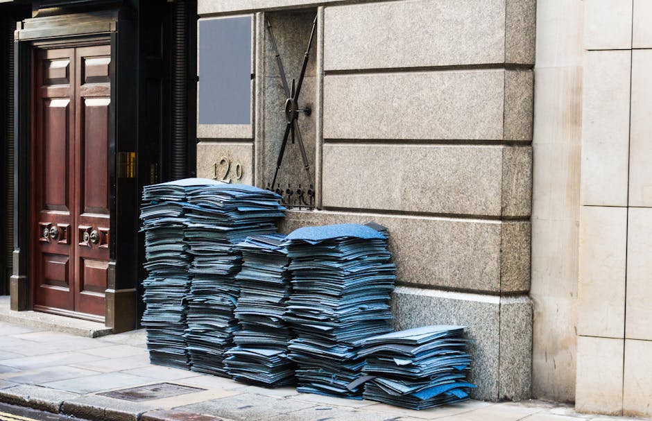 A large pile of blue cardboard folders and document boxes stacked against a stone building exterior, positioned on the pavement outside a residential property. The documents are organized in multiple stacks, with some leaning slightly. To the left, there are dark wooden doors with decorative panels, and the building features a metallic wall-mounted object, possibly a decorative gate or fixture, adjacent to the boxes. The scene is illuminated by natural daylight, providing clear visibility of the materials and environment. This setup suggests preparation for house removals or packing, indicative of furniture transport or packing and moving activities, with the stacks representing documents or packaging materials ready for removal by a professional service like Marylebone Man and Van, specialists in house removals and relocation services.