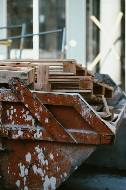 A close-up view of a rusty, weathered boat used as a makeshift mobile storage or staging area outside a residential property. On the boat, there are several wooden crates and pallets, some of which are reinforced with metal straps, possibly for moving or packing purposes. The crates are made of light-colored wood, with some showing signs of wear, and are positioned towards the front end of the boat, ready for loading or transport. The boat is situated on a paved surface near the entrance of a house, with a partially visible glass door and window in the background, along with construction materials and scaffolding indicating ongoing building or renovation work. Inside the boat, a few loose cardboard boxes and packing materials can be seen, suggesting preparation for a home relocation or furniture transport process. Occasional dust and paint splatters are visible on the boat's exterior, which is typical for outdoor storage during moving activities. The scene reflects an organized setup for loading household items, with a focus on furniture transport and packing logistics involved in a professional removal service like those offered by Marylebone Man and Van.