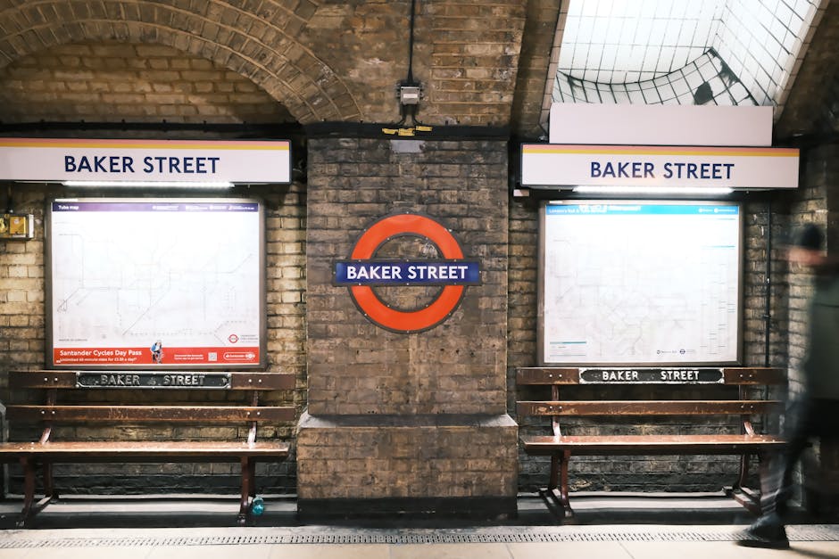 The image depicts the underground Baker Street station in London, featuring two large illuminated station maps behind wooden benches placed against a brick wall. The maps are enclosed in protective covers and display the rail network, with the station name 'BAKER STREET' prominently shown on signs above each map. A round, red London Underground logo is mounted on the central brick pillar between the two maps. A person in dark clothing, blurred due to motion, is walking past the right side of the frame. The station environment is lit with artificial lighting, highlighting the brickwork and fixtures, creating a typical underground transit setting. This scene is relevant to home relocation and furniture transport services provided by Marylebone Man and Van, illustrating the urban underground infrastructure often involved in moving logistics or access considerations for house removals near the Sherlock Holmes Museum area in Marylebone.