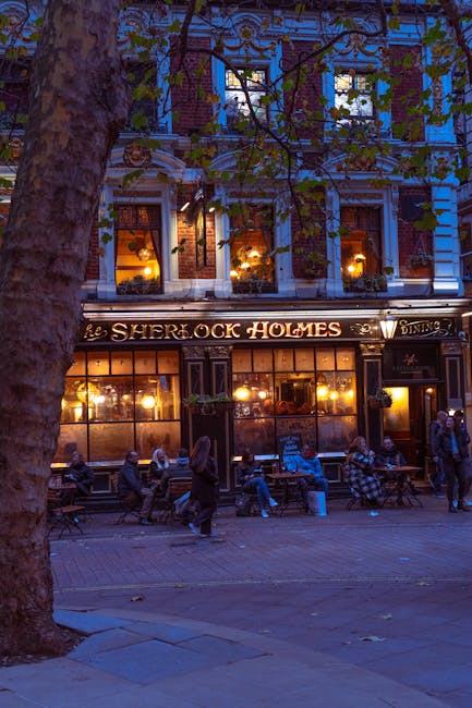 A street scene outside a traditional pub with illuminated signage reading 'The Sherlock Holmes', featuring large glass windows displaying warm interior lighting. The multi-storey building has ornate architectural details and is adorned with hanging flower baskets and decorative lighting. In front, several people are sitting on benches and street furniture, some engaged in conversation or using phones, while others stand nearby. The area is shaded by a large tree with green leaves, and a few individuals are walking along the pavement, which is suitable for home relocation or furniture transport activities, as managed by Marylebone Man and Van, a specialist in removals and moving services.