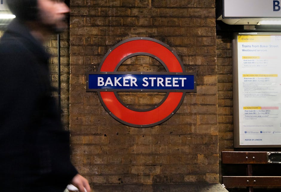 The image depicts the underground Baker Street station in London, featuring two large illuminated station maps behind wooden benches placed against a brick wall. The maps are enclosed in protective covers and display the rail network, with the station name 'BAKER STREET' prominently shown on signs above each map. A round, red London Underground logo is mounted on the central brick pillar between the two maps. A person in dark clothing, blurred due to motion, is walking past the right side of the frame. The station environment is lit with artificial lighting, highlighting the brickwork and fixtures, creating a typical underground transit setting. This scene is relevant to home relocation and furniture transport services provided by Marylebone Man and Van, illustrating the urban underground infrastructure often involved in moving logistics or access considerations for house removals near the Sherlock Holmes Museum area in Marylebone.