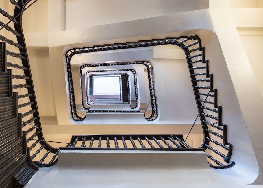 A view looking down a vertical staircase inside a building, with multiple landings and black metal handrails on each side, forming a square spiral pattern that descends towards a rectangular opening at the bottom. The staircase walls are painted white, and natural light filters in from a window visible at the bottom, illuminating the stairwell. The image captures the structural details and the enclosed environment suitable for professional moving and furniture transport services, such as those offered by Marylebone Man and Van, especially during home relocation or packing and moving processes.