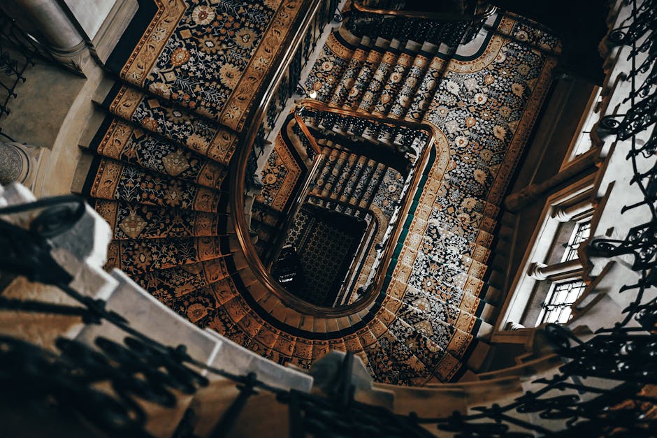 A view looking down a winding staircase inside a property on Wimpole Street, featuring a detailed, patterned carpet runner on each step and wooden handrails on either side supported by decorative wrought iron balustrades. Natural light filters through nearby windows, illuminating the ornate interior. The staircase descends from the top floor, revealing a series of curved steps with their edges covered by protective padding. At the bottom, a section of the patterned carpet extends across the landing, with a glimpse of a doorway and wall partially visible. This interior scene captures the intricate architectural elements and traditional decor, typical of a historic or period building in Marylebone. The image is part of a house relocation or moving process conducted by Marylebone Man and Van, who are handling furniture transport and heavy-item moving within the property, involving careful handling of furnishings, packing materials, and moving equipment visible near the staircase.
