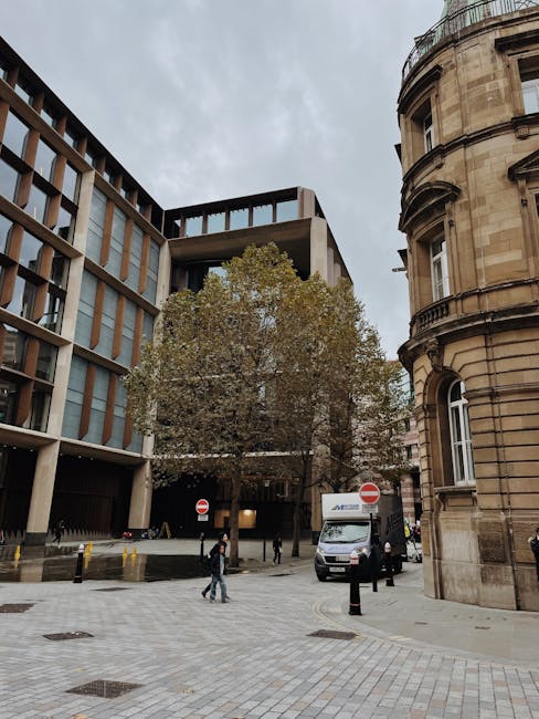 A street scene in Marylebone, showing a modern multi-storey office building with large windows on the left and a historic, rounded stone building with arched windows on the right. In the foreground, a grey van with the Marylebone Man and Van logo is parked next to the pavement, loading a large cardboard box wrapped in plastic. Two workers, dressed in casual clothing, are lifting a piece of furniture or box onto or off the van, with a dolly and other packing materials visible nearby. Several pedestrians walk across the paved street area, which is marked with black and yellow bollards, while a large tree with sparse leaves stands between the buildings. The scene is set during daytime under an overcast sky, capturing the typical urban environment involved in home relocation or furniture transport, as part of moving services connected to the W1U postcode and parking/loading considerations.