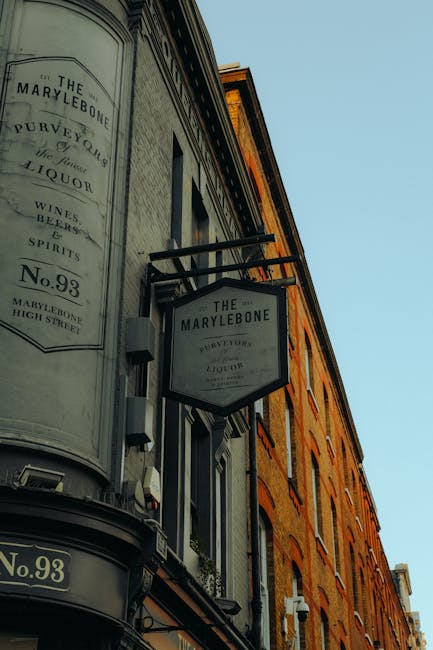 A photograph captures the exterior of a multi-storey building in the Marylebone area, featuring signage for 'The Marylebone' pub or shop, mounted beneath a small overhang. The building's façade is constructed of red brick, with multiple windows visible along its height. The signage includes a large rectangular sign and a smaller octagonal one, both displaying the establishment name. The image is taken from a low angle, showing clear daylight and blue sky above. This environment suggests a typical London street scene, where local business signage and buildings are visible, relevant to house removals and relocation services such as those offered by Marylebone Man and Van, supporting the context of home and furniture transport or moving logistics.